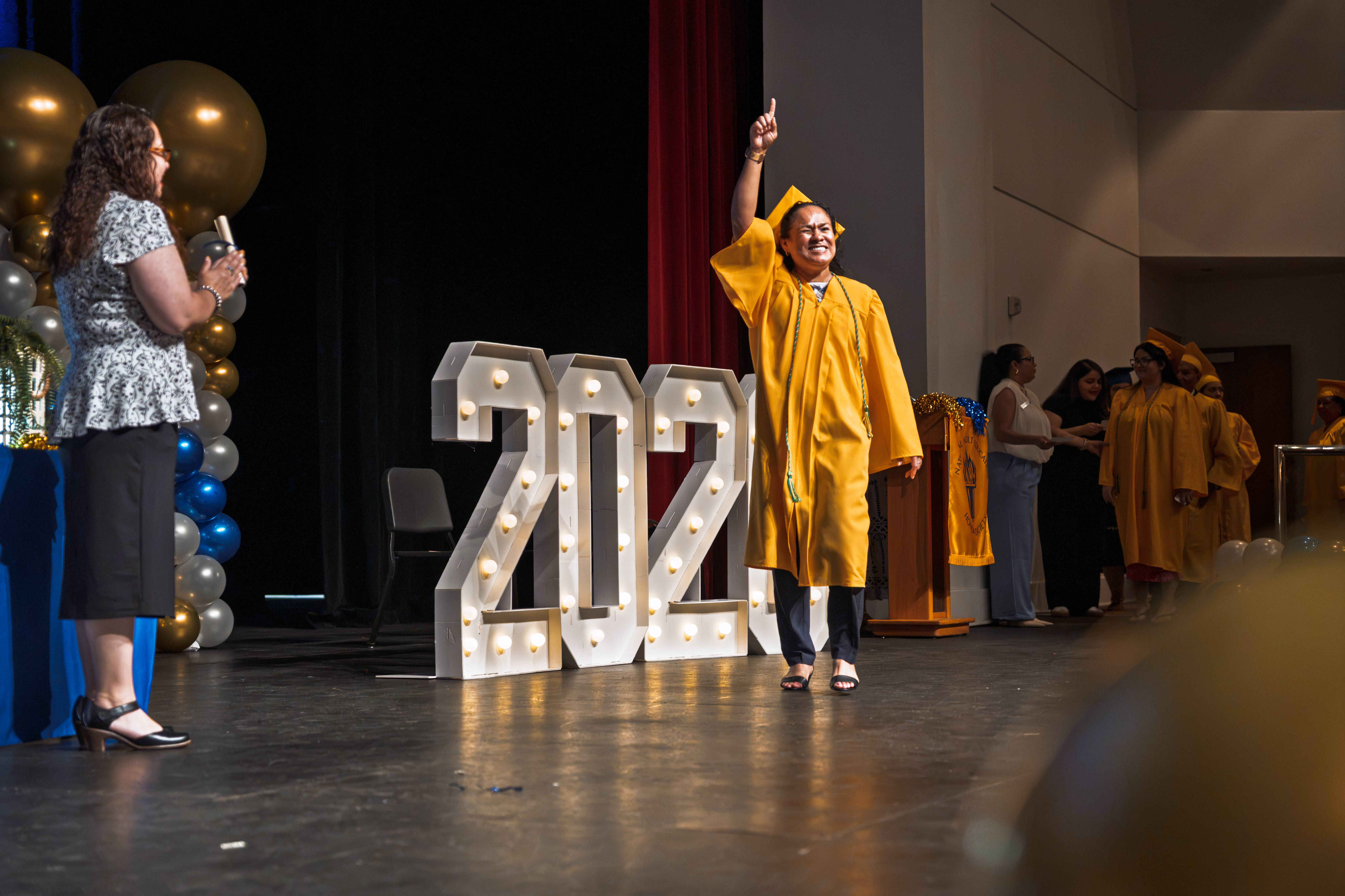 A graduate in cap and gown points skyward while crossing the stage beside the lighted 2026 sign