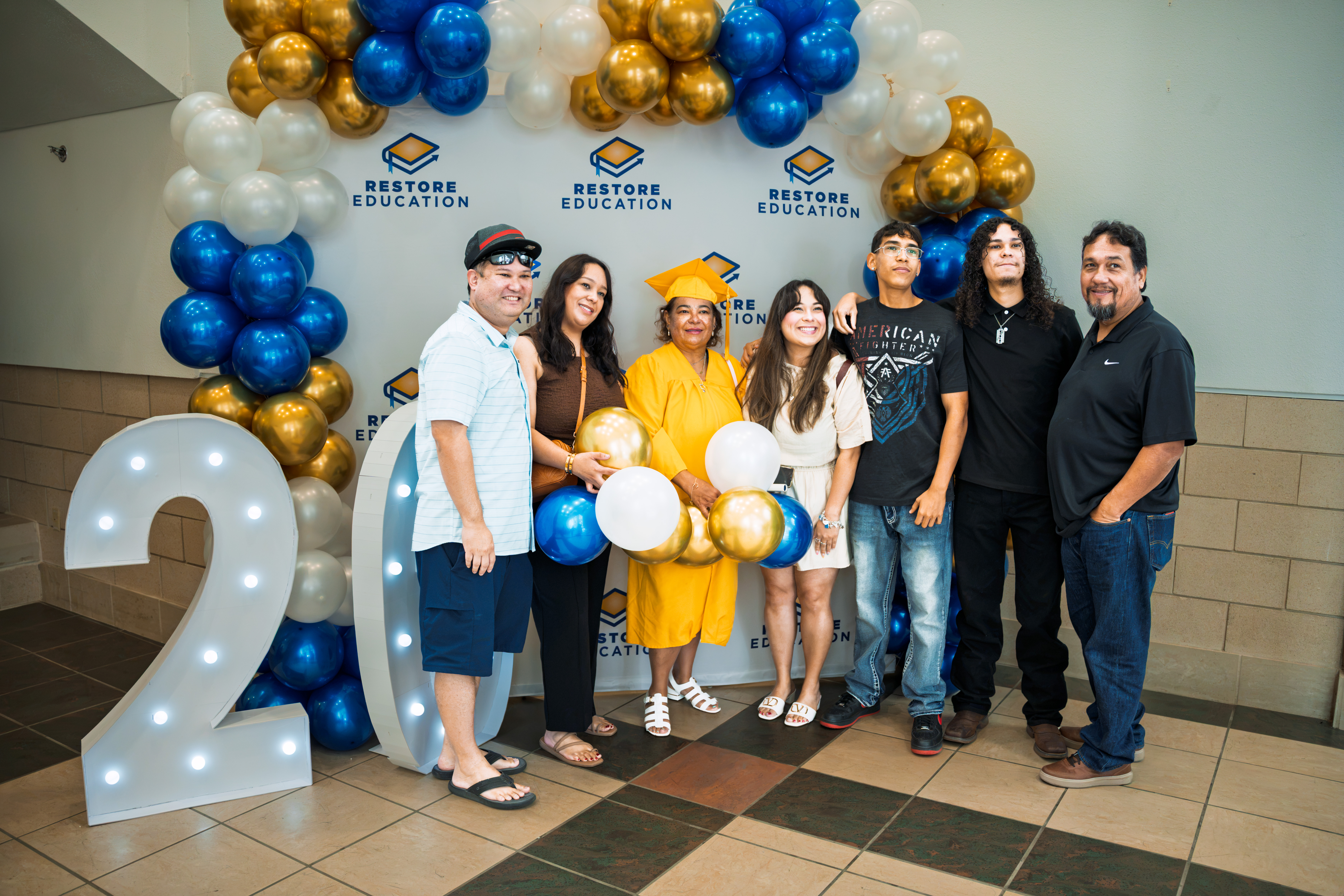 A graduate in a gold gown poses with her family in front of the Restore Education balloon backdrop